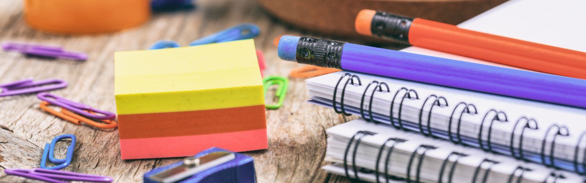 eine bunte Auswahl an Büro- und Schulmaterialien wie Notizblöcke, Haftnotizen, Bleistifte, Büroklammern und einen Spitzer auf einem Holztisch